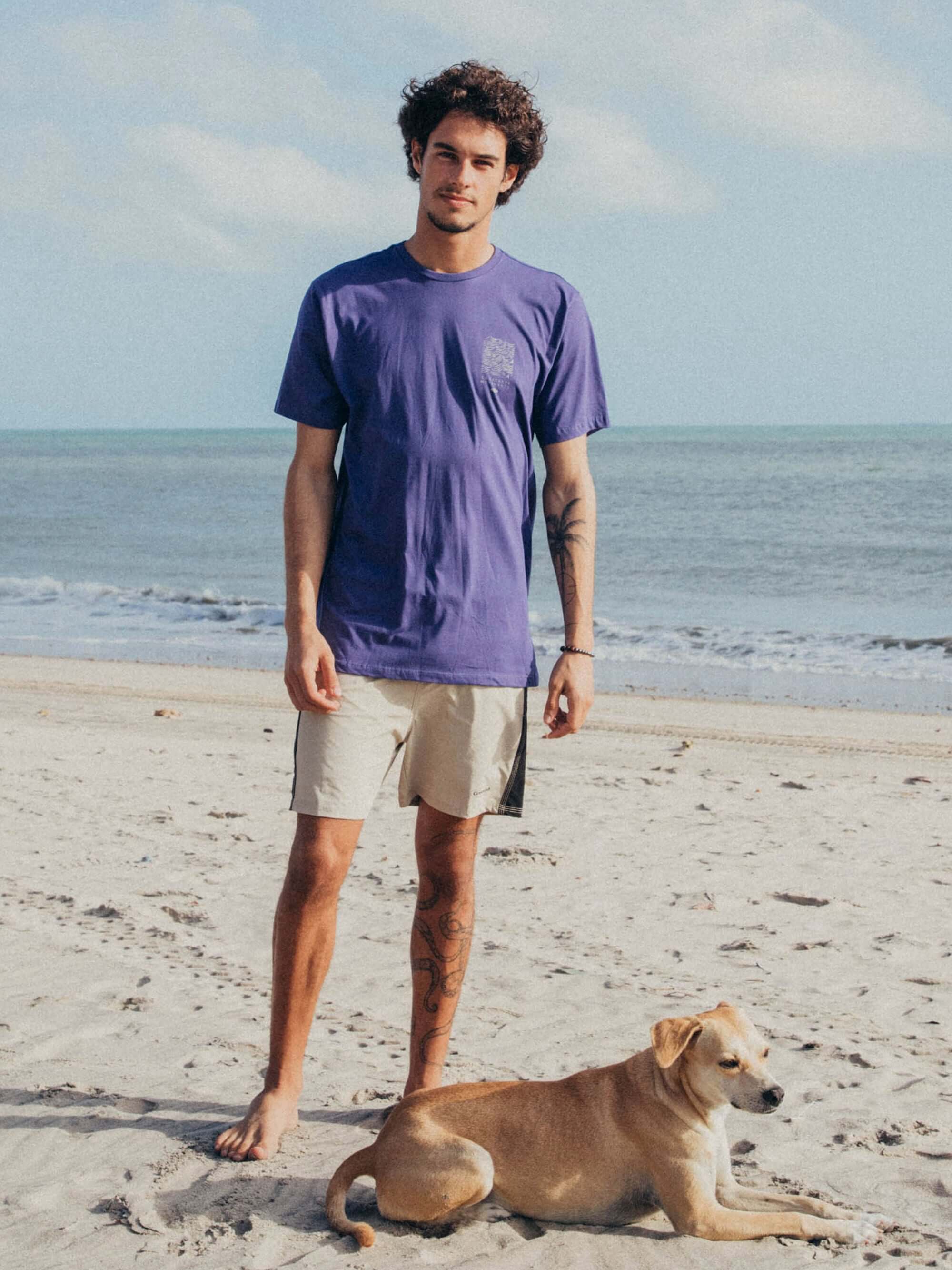 Homem na praia usando camiseta roxa da Greenish e short claro, com cachorro ao seu lado.