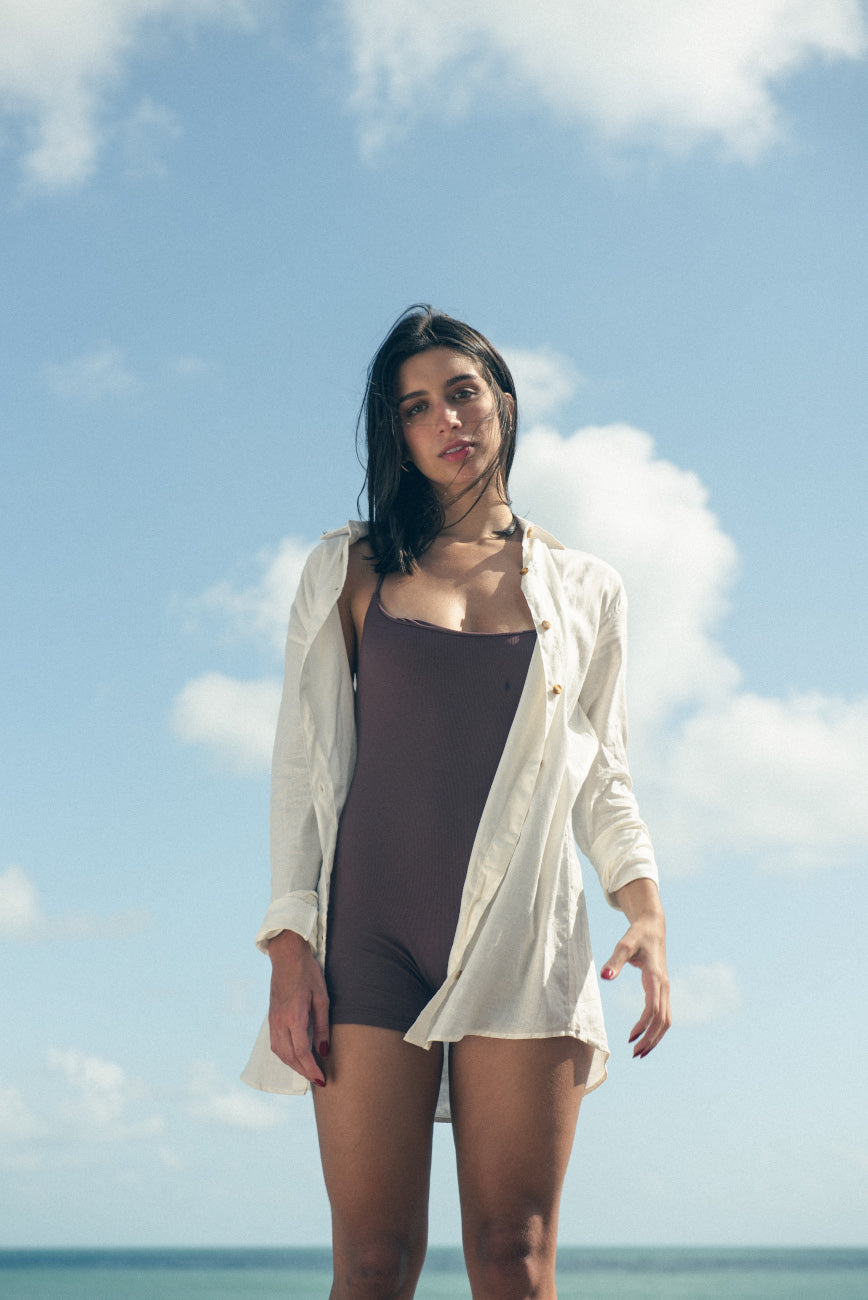 Modelo feminina em maiô marrom e camisa branca, posando contra o céu azul e nuvens.