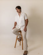 Man wearing Greenish Camisa Botao High Tide - Off White sitting on stool in light background, showcasing casual style.