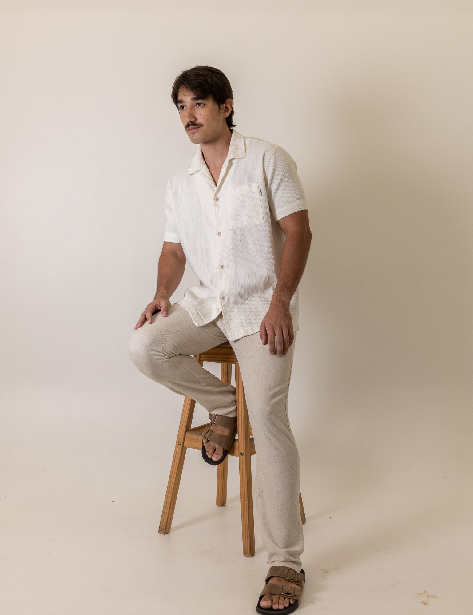 Man wearing Greenish Camisa Botao High Tide - Off White sitting on stool in light background, showcasing casual style.