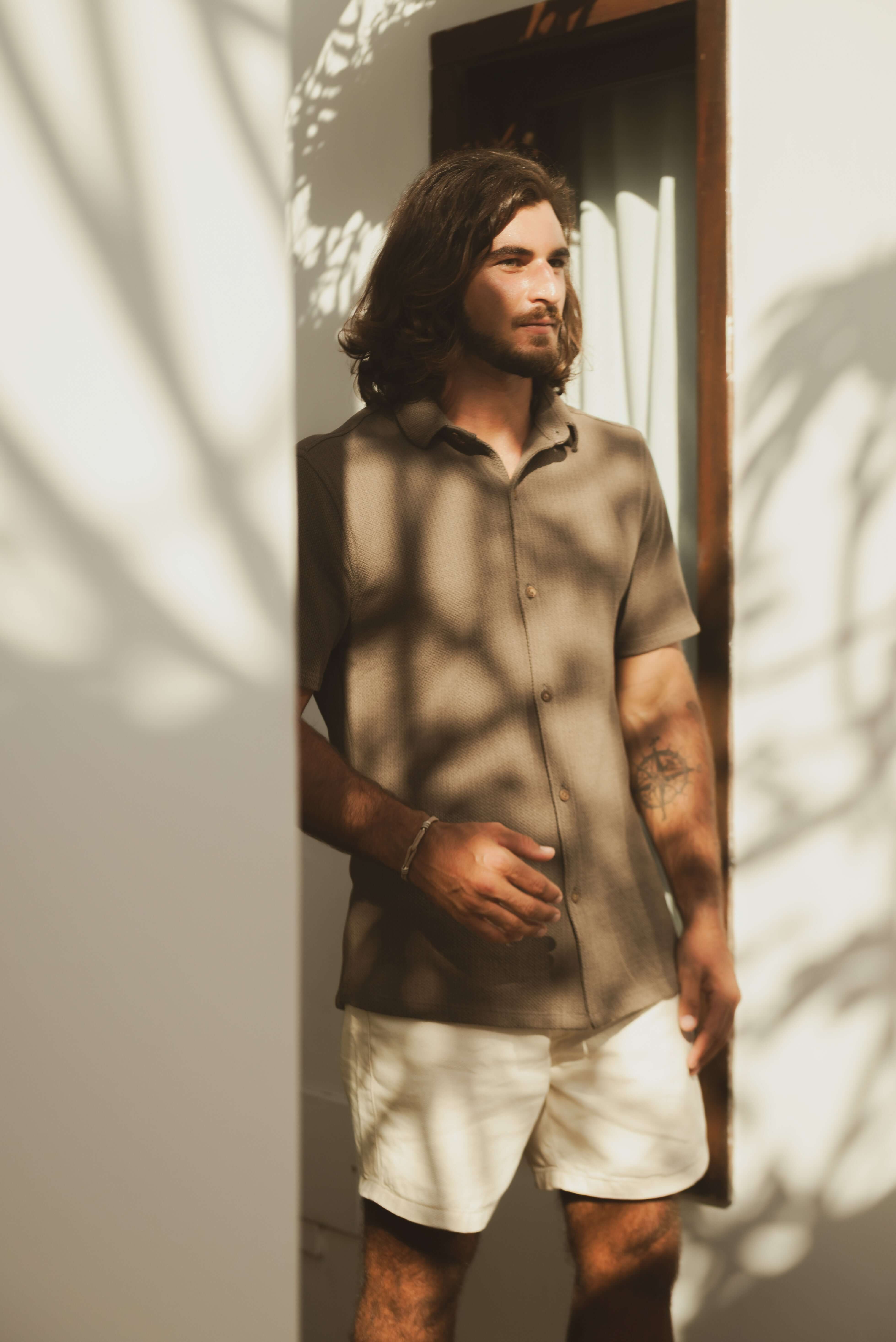 Homem usando Camisa Botão Maré - Verde Escuro, destacando elegância casual e conforto em um ambiente iluminado por sombras de folhas.