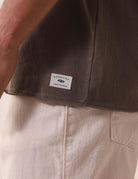 Detalhe da REGATA LARGA WAVES - VERDE ESCURO da marca Greenish com etiqueta na base, destacando a qualidade e estilo em algodão 100%.