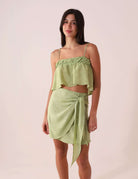 Modelo usa a SAIA CURTA THÁ - VERDE por Greenish, destacando amarração lateral e sobreposição. Saia em viscose macia e leve.