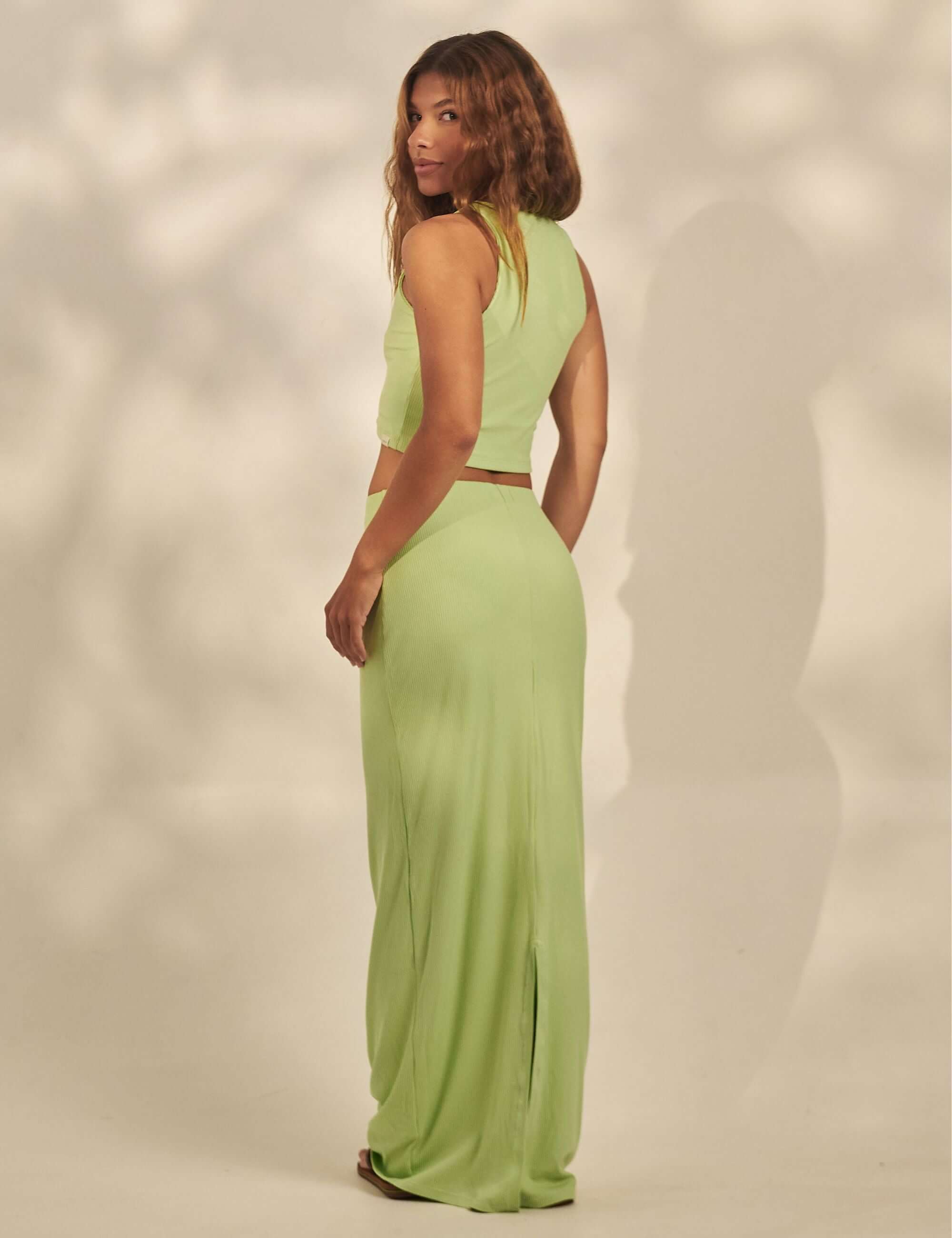 Saia Longa Canelada Emma - Verde Claro da Greenish mostrada em modelo de costas, tecido canelado com fenda e caimento fluido