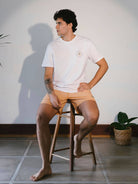 Modelo usando camiseta Roots Native branca da Greenish, sentado em um banco de madeira, com estilo casual.