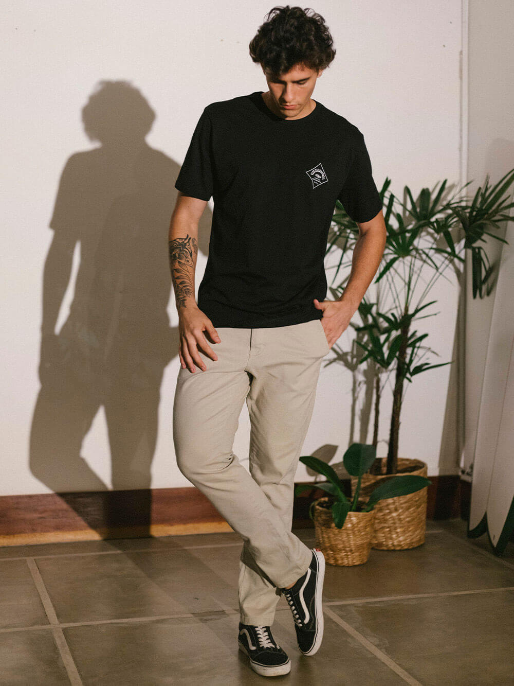 Camiseta Hibiscus e Planks da Greenish em preto, modelo masculino em ambiente casual com plantas decorativas.