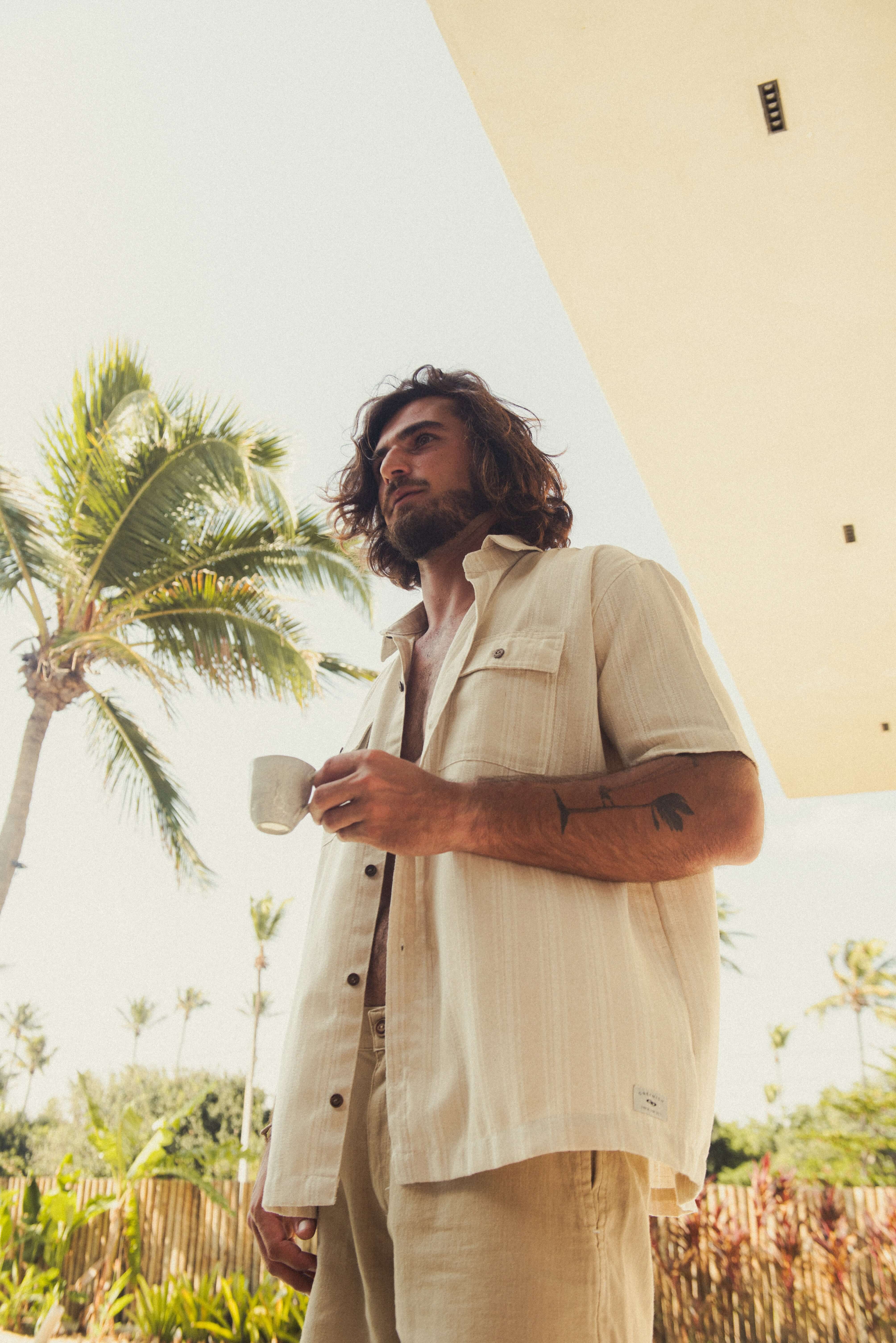 Homem com Camisa Botão Linho Listrado Oversized - Natural da marca Greenish, em cenário tropical ao ar livre. Moda utilitária sofisticada.