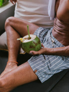 Homem relaxando em sofá com coco verde na mão, usando short listrado da marca Greenish.