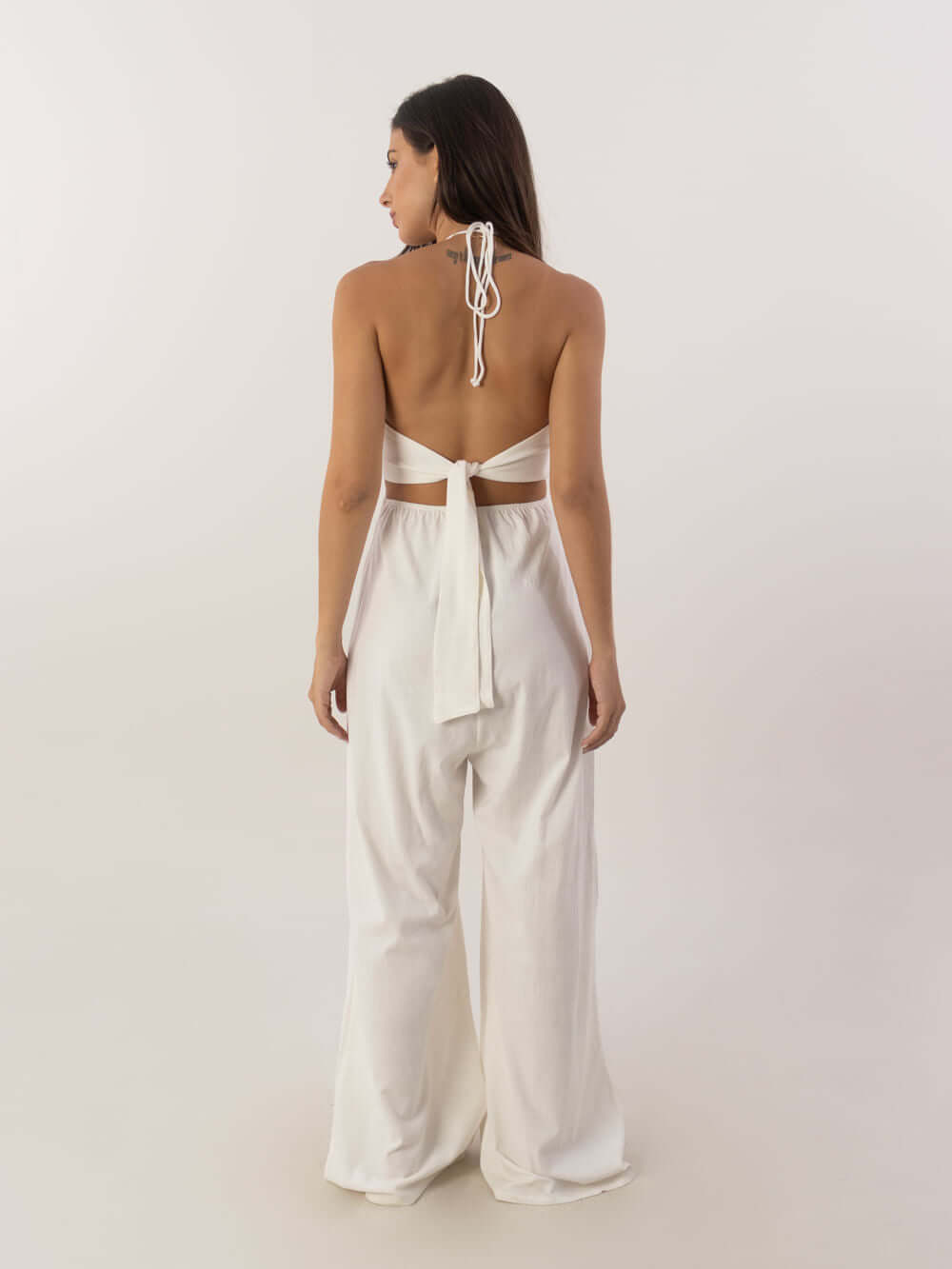 Mulher mostrando o macacão longo Alyssa off white da Greenish, com decote em V e amarração nas costas.