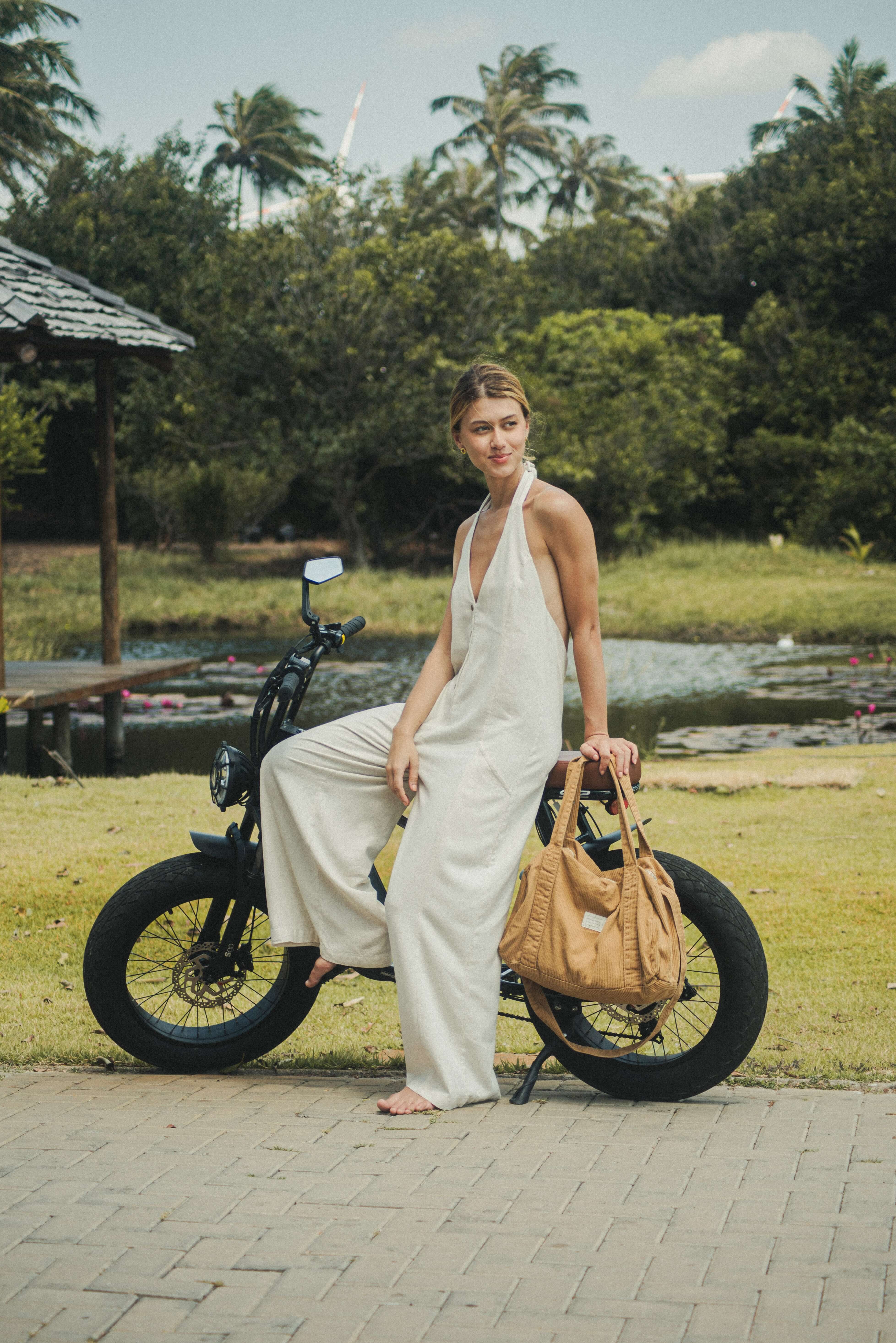 Mulher usando o Macacão Longo Gaia - Natural da Greenish, tecido de linho leve e fresco, em ambiente natural ao lado de uma bicicleta elétrica.