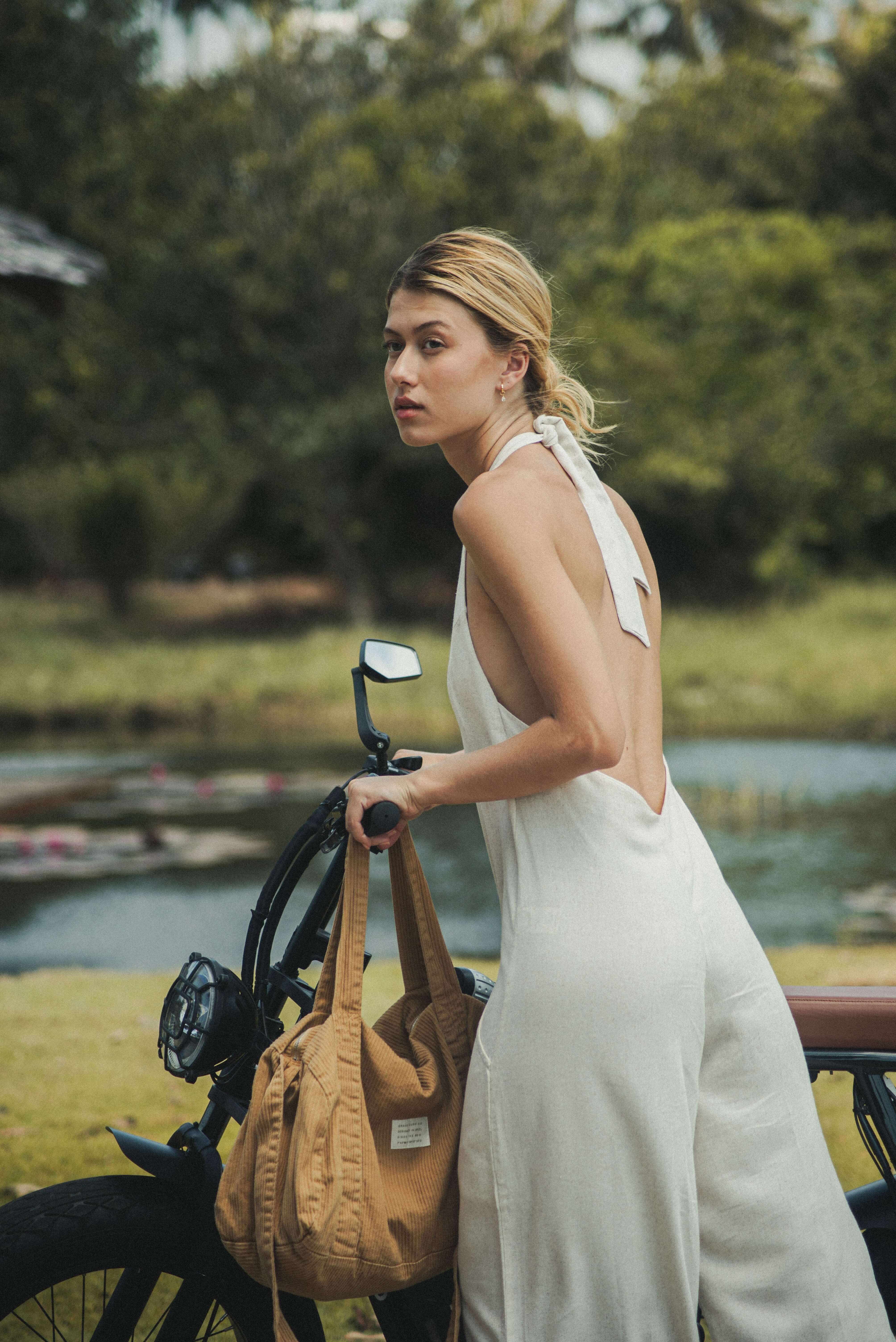 Mulher usando Macacão Longo Gaia - Natural da Greenish, em linho leve, com decote halter e amarração, ao lado de uma bicicleta em ambiente natural.