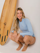 Modelo usando o Maiô Greenish Bayshore azul, posando ao lado de uma prancha de surf, destacando conforto e estilo para práticas aquáticas.