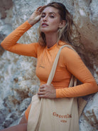 Stylish woman in an orange Bayshore swimsuit holding a tote bag against a rocky background.