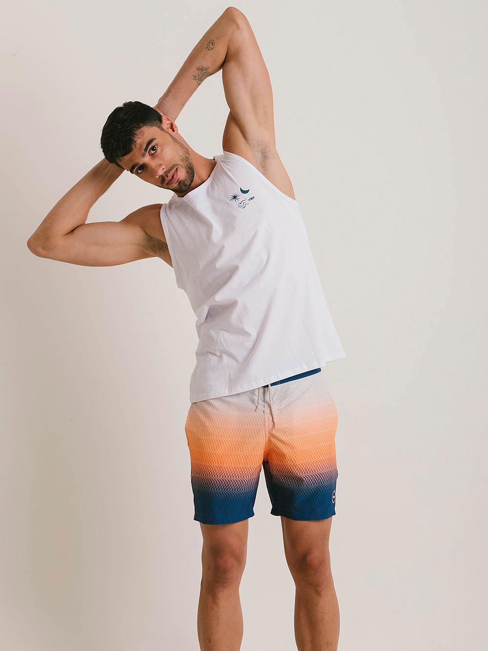 Homem fazendo alongamento com regata branca da marca Greenish e bermuda com degradê em tons de laranja e azul.