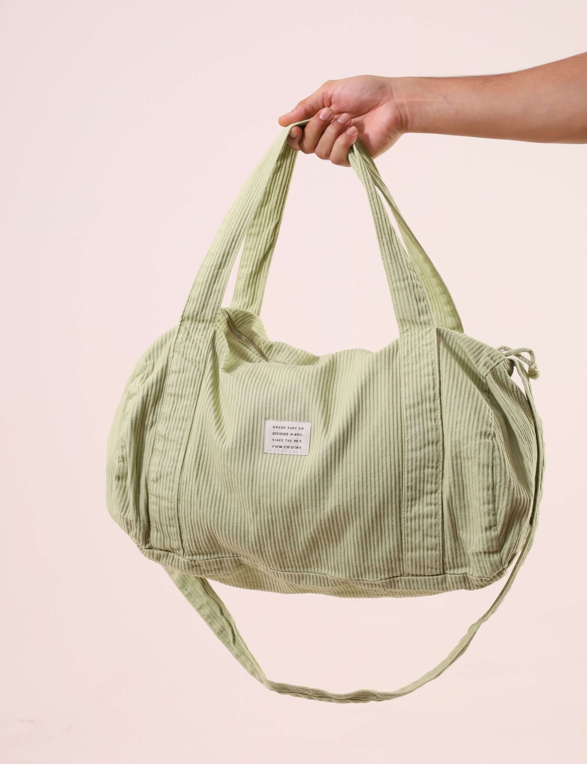 Hand holding a Sacola Duffel Bag - Verde Claro, showcasing its reinforced fabric handles and soft textured peletizada canvas in light green.