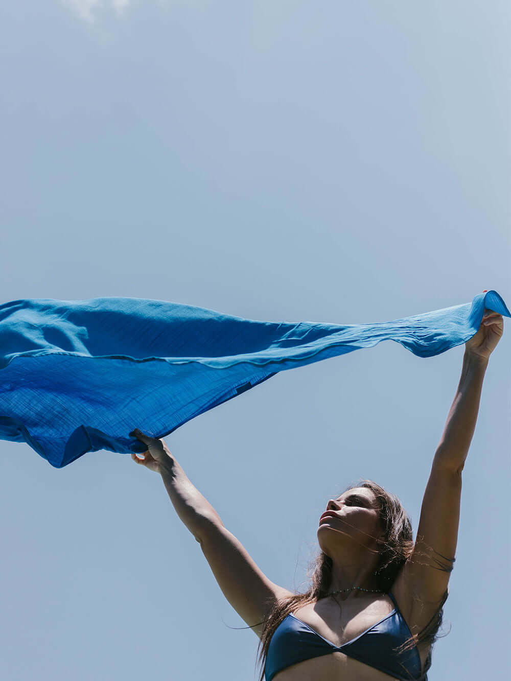 Mulher segurando sarong azul Praia Lana da marca Greenish ao ar livre, sob o céu claro.