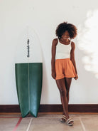 Modelo usando shorts laranja da Greenish ao lado de uma prancha de surf, estilo casual e descontraído.