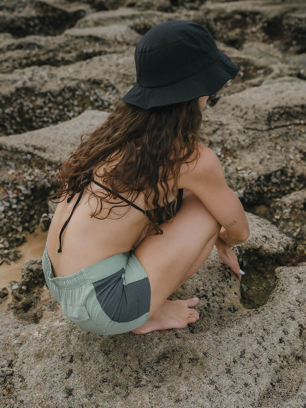 Modelo usando Short Sport Sidecut Verde da Greenish, explorando rochas na praia com estilo e conforto.