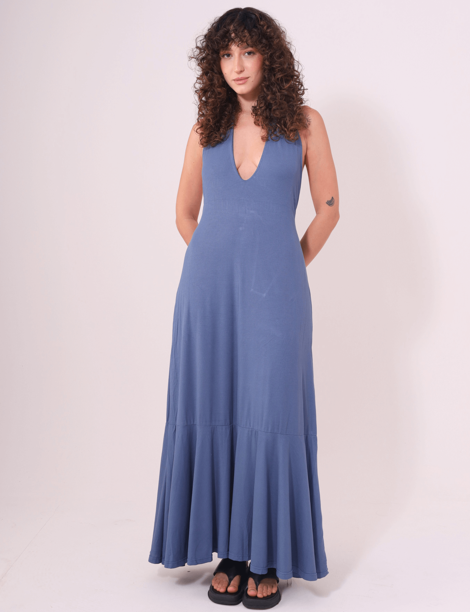 Mulher usando Vestido Longo Serena Azul Jeans com decote profundo e jabô na barra, da marca Greenish, ideal para dias de calor.